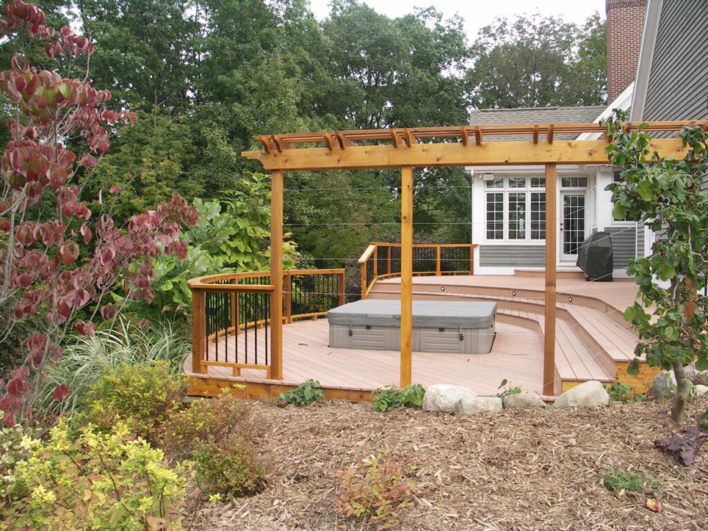 A pergola and deck setup with a curved layout design to fit a hot tub inset. For the best deck builders in Rockford MI has to offer, call Signature Decks & Construction.