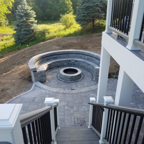 An outdoor fire feature (fire pit) being built in the middle of a circular stone patio set, steps away from the steps of an upper-floor deck.